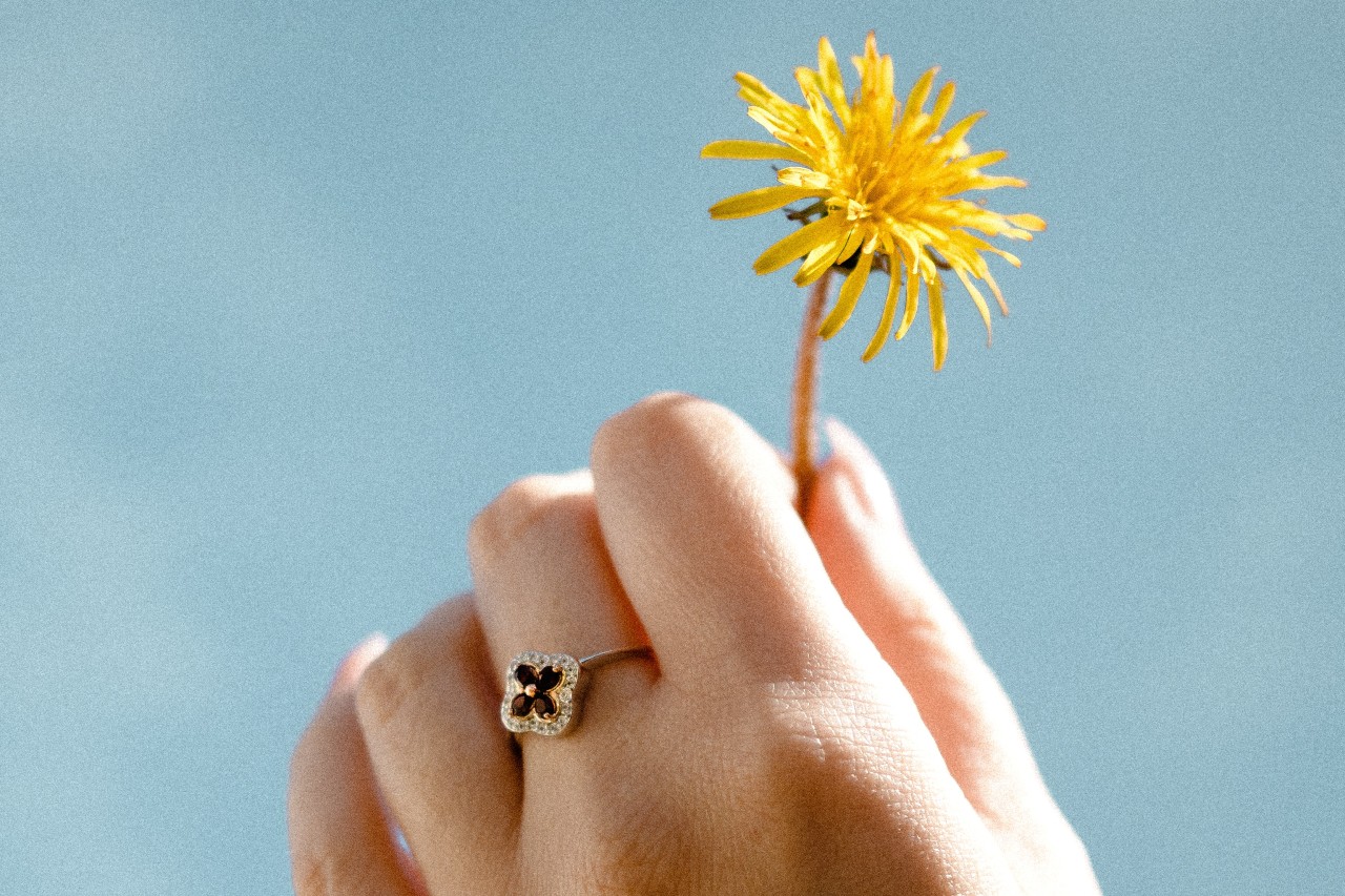 Close up image of a hand holding a dandelion and wearing a dainty ring with a flower motif