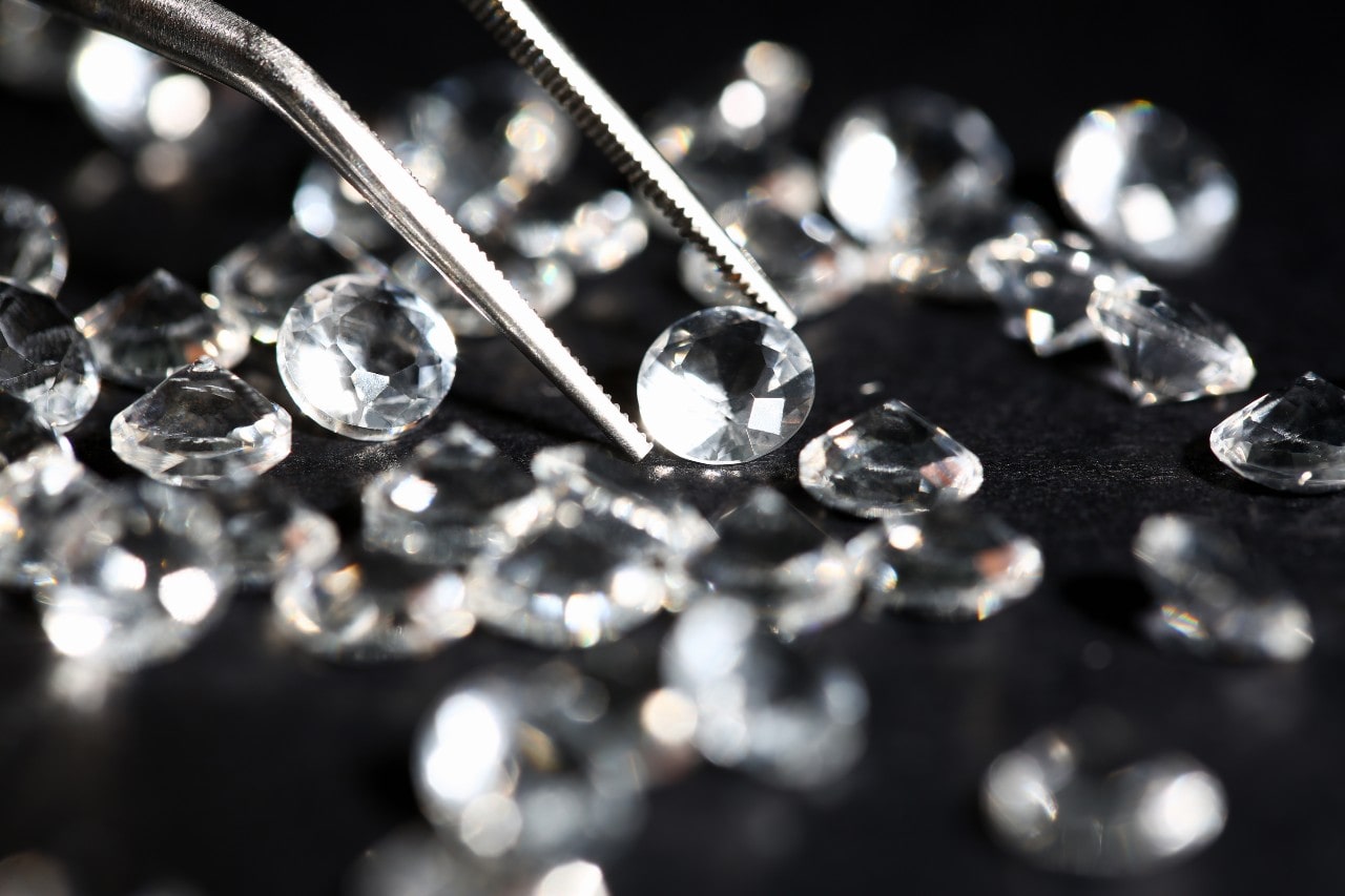 A round cut diamond sits in a pair of jeweler tweezers among a bunch of other diamonds on a black background