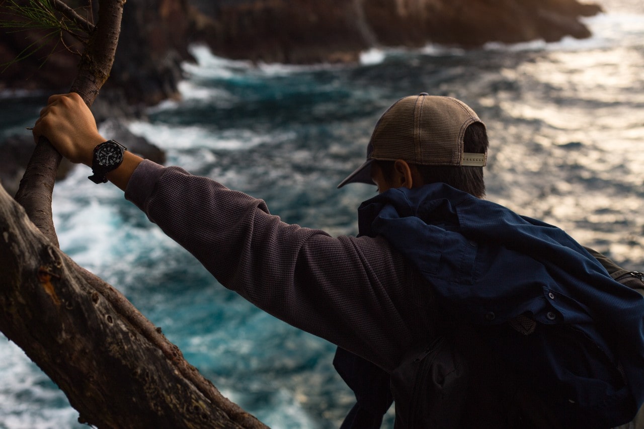 A hiking man leans against a tree and watches a river flow while wearing a watch