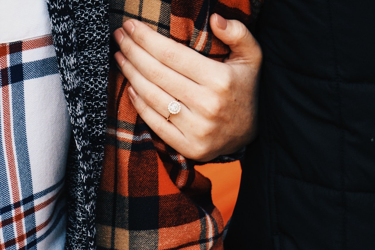 A woman’s hand holding her partner’s arm and wearing a rose gold, halo engagement ring