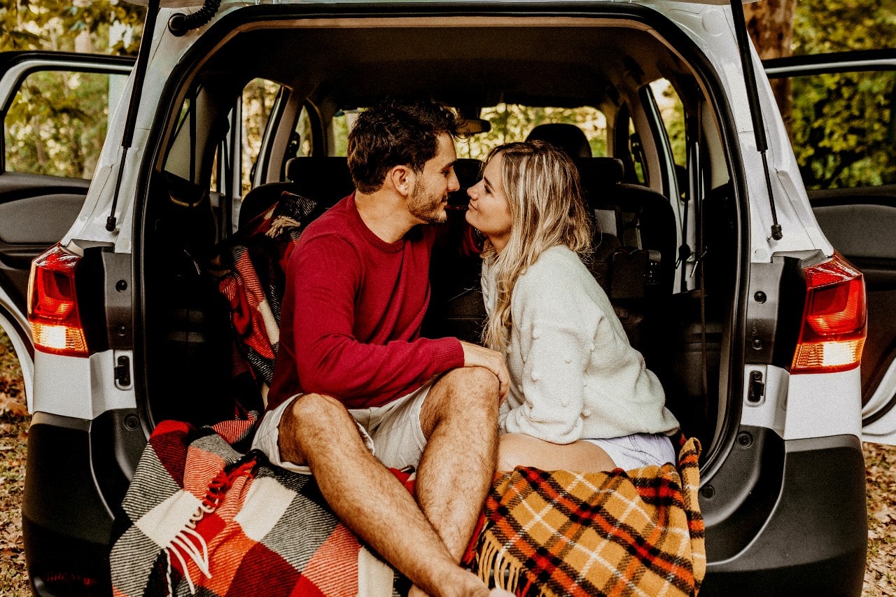 A couple sitting in the back of an SUV, smiling at one another and wearing fall colors