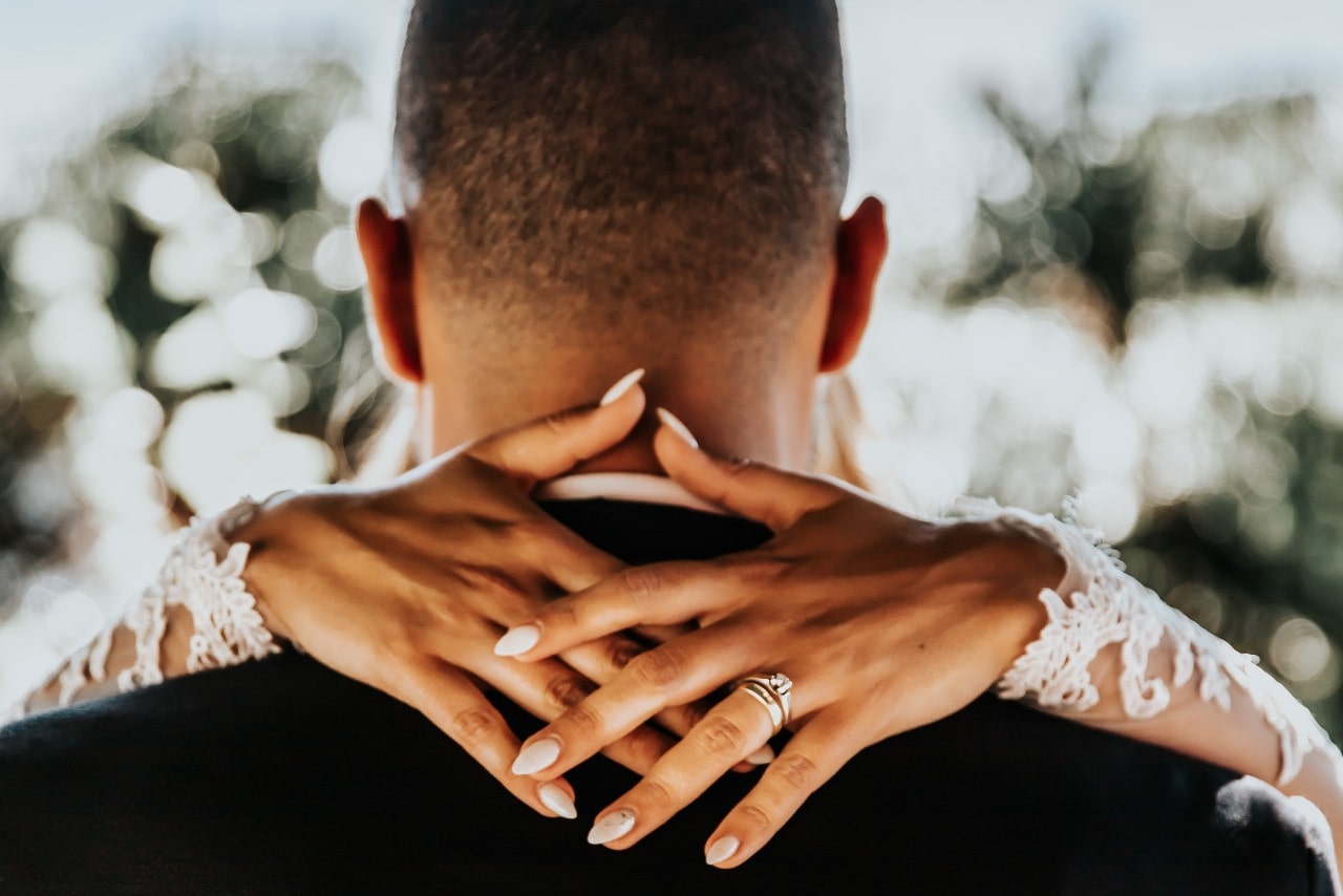 A bride rests her hands around her groom, showing off her rings.