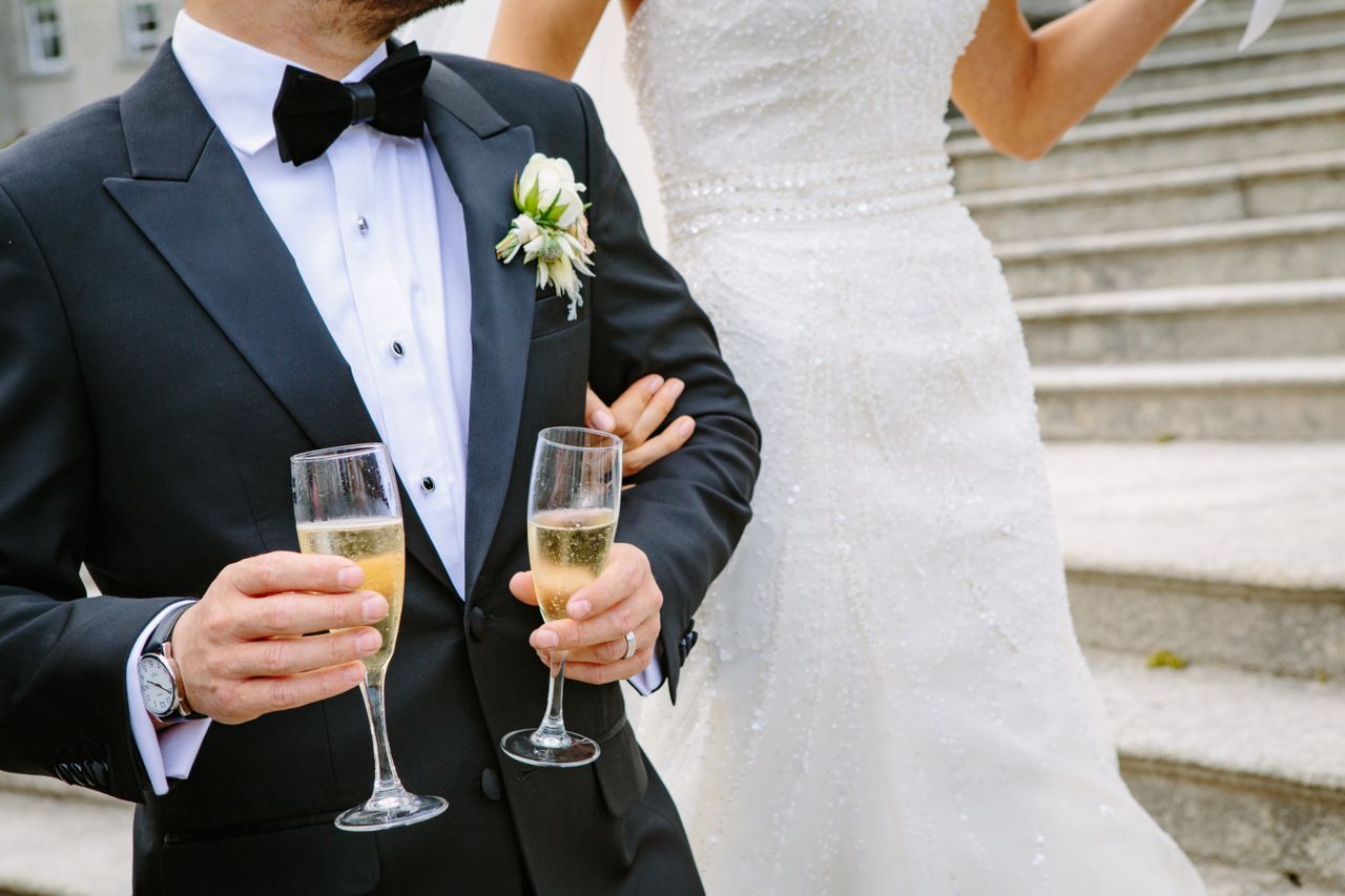 A groom holds two glasses of champagne as his bride is on his arm.