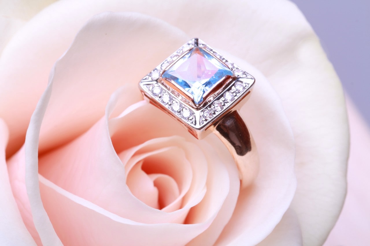 A square-cut gemstone ring with a sparkling diamond border set on a rose gold band, elegantly displayed on a soft pink rose.