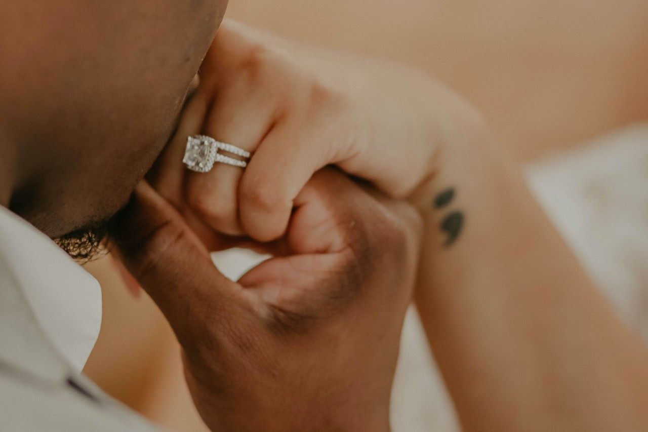 A man kissing his bride-to-be’s hand, a cushion cut engagement ring on her finger.