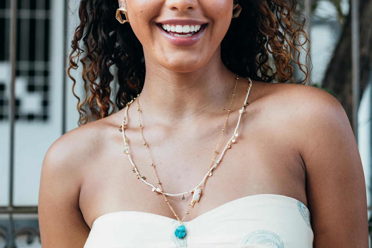 Woman with curly hair wearing a layered yellow gold necklace featuring a blue gemstone at the center. Woman with curly hair wearing a layered yellow gold necklace featuring a blue gemstone at the center.