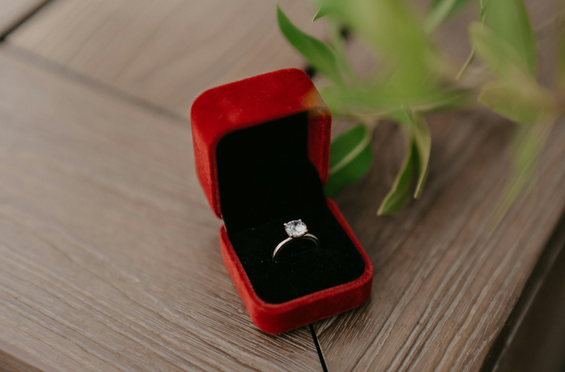 A red ring box sitting on a wooden table and holding a solitaire engagement ring.