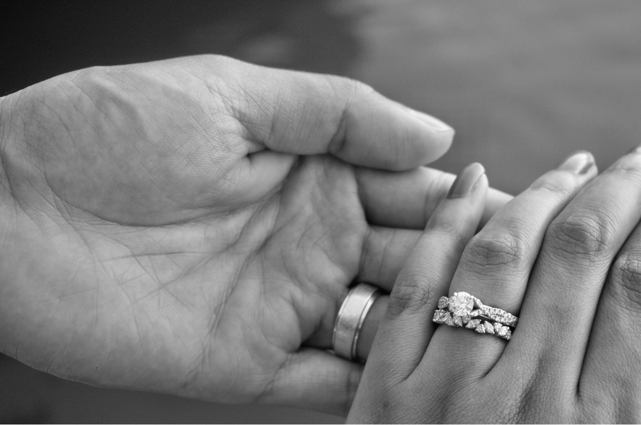 A black and white photo of a man’s hand holding a woman’s that is adorned with a diamond engagement ring.