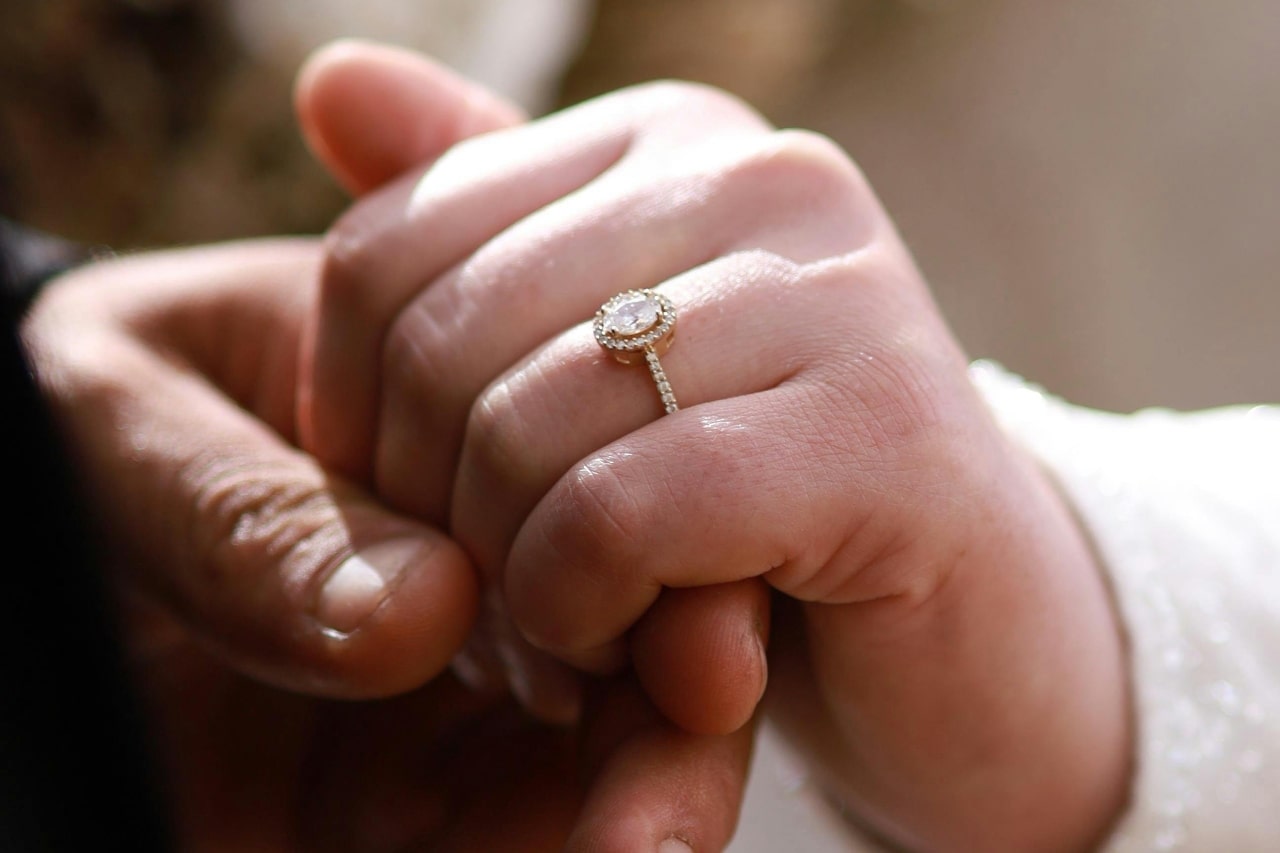 A man’s hand holding a woman’s that is adorned with an oval cut halo engagement ring.