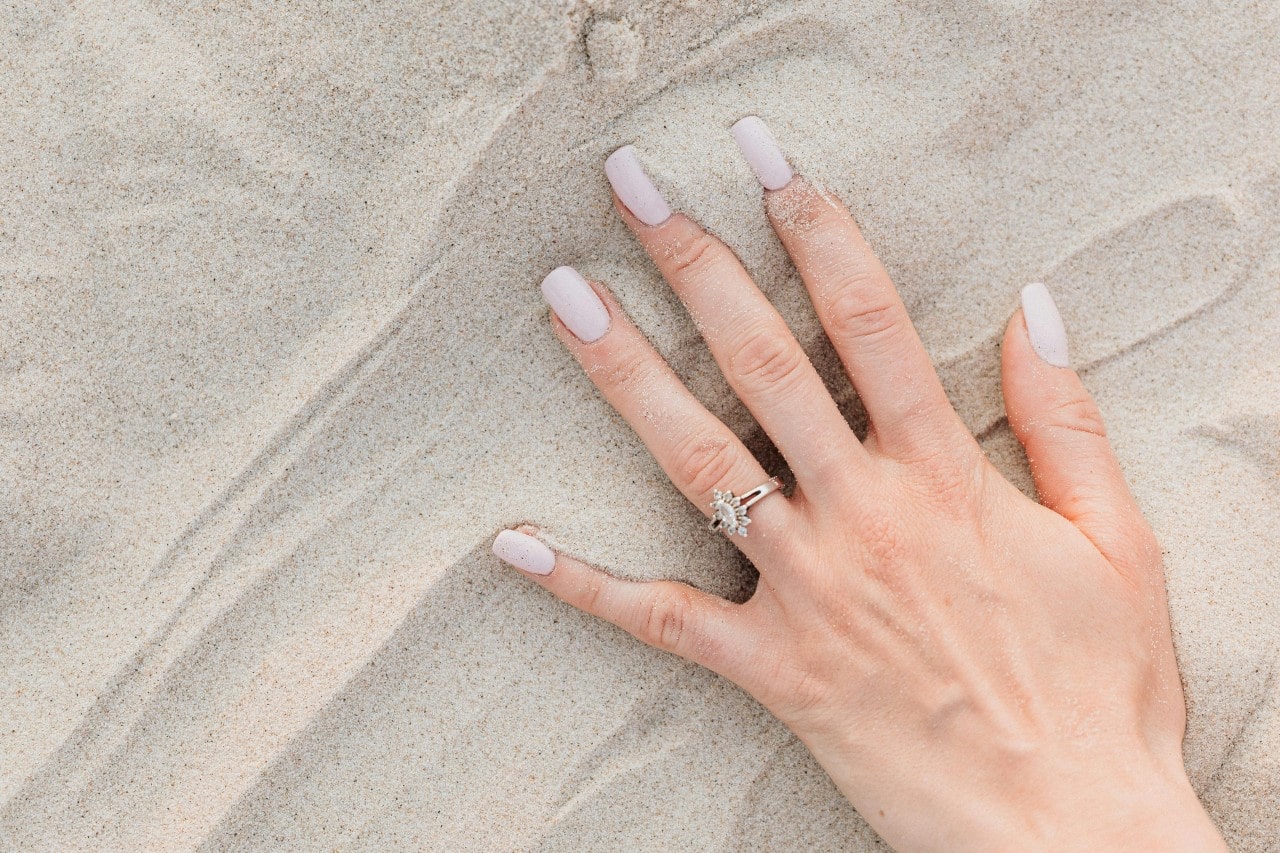 a woman’s hand in the sand, wearing a unique halo engagement ring